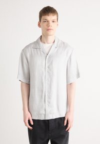 Camicia grigio chiaro a maniche corte con colletto a punta, chiusura frontale con bottoni e vestibilità comoda, abbinata a pantaloni scuri. Tessuto dalla texture liscia.