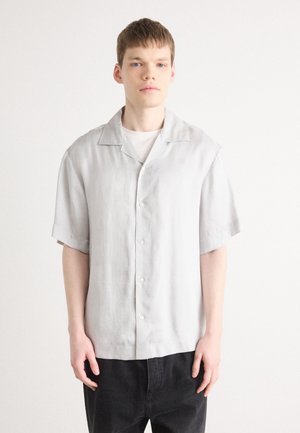 Camisa de manga corta en gris claro con collar puntiagudo, cierre frontal con botones y un corte relajado, combinado con pantalones oscuros. Textura de tela suave.