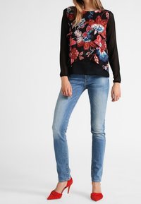 Blouse à motifs floraux avec manches noires, design superposé ; assortie à un jean bleu clair et des chaussures à talons rouges.