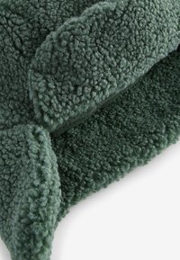 Zelená plyšová fleece tkanina s textúrovaným, chlpatým povrchom. Obsahuje ohnutý okraj a mäkkú, hrubú štruktúru pre teplo a pohodlie.
