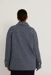 Manteau à motifs vichy bleu et noir avec un large col, de gros boutons et une coupe structurée, comportant des manches longues et un tissu texturé.