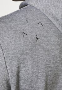 Šedá mikina s kapucí vyrobená z texturované tkaniny, s černým logem a rozptýleným nápisem na zadní straně. Minimalistický design s pevným límcem.