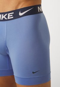 Plave Nike bokserice s tamnoplavim pojasom na kojem se nalazi bijelo slovo "NIKE". Glatka tekstura, uski kroj i crni logo detalj na nozi.