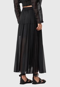 Gonna plissettata nera in tessuto trasparente, con vita elastica e lunghezza alla caviglia. Abbinata a sandali neri piatti con dettagli dorati.