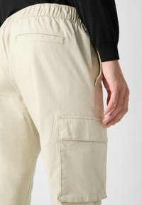 Beige Cargo-Hosen mit einem elastischen Bund, die seitliche Taschen und eine Gesäßtasche haben. Der Stoff hat eine glatte, leicht gerippte Textur.