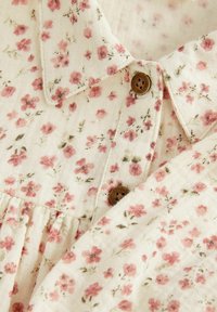 Blouse à motif floral avec un fond crème orné de fleurs roses, un col et deux boutons marron pour la fermeture. Tissu texturé avec des plis.