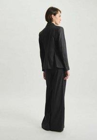 Costume pour femmes sur mesure en gris foncé avec des rayures verticales. Le blazer présente des revers crantés ; le pantalon est à jambe large avec une coupe droite.