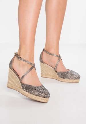 Pieds portant des espadrilles compensées argentées ornées de strass avec brides à la cheville sur un fond neutre.