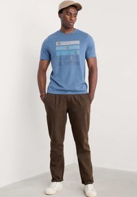 En blå bomullströja med en grafisk design av en måne och ränder, ihopparad med bruna texturerade byxor och vita sneakers.