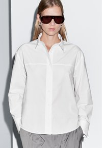 Donna che indossa una camicia bianca oversize con bottoni, grandi occhiali da sole scuri, orecchini a cerchio argentati e pantaloni grigi plissettati su sfondo semplice.