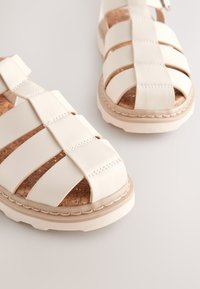 Sandales blanches en cuir avec semelles intérieures en liège, dotées de larges sangles croisées et d'une semelle rembourrée. Des détails de couture ajoutent de la texture.