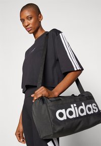 Μαύρο σακίδιο duffel με λευκό λογότυπο "adidas", κατασκευασμένο από ανθεκτικό υλικό, με φερμουάρ και ανθεκτικούς ιμάντες ώμου.