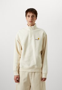 Sweatshirt zippé beige clair avec un col montant, des poignets côtelés et un petit logo brodé sur la poitrine. Texture douce et coupe décontractée.