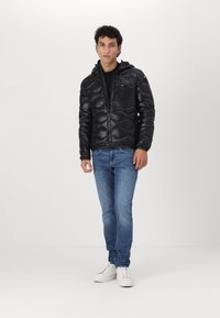 Giacca a vento nera con design trapuntato e cappuccio, dotata di una cerniera frontale e una tasca laterale. Indossata con jeans blu e sneakers bianche.