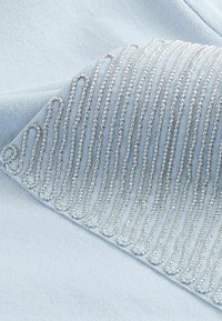 Vaaleansininen kangas esittelee koristeellista hopeista helmi-uurretta, joka muodostaa kiertäviä viivoja ja luo siten tekstuuriltaan kerroksellisen ilmeen.