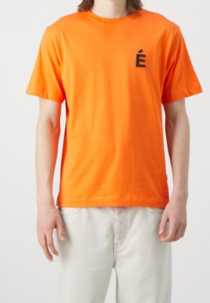 Personne portant un t-shirt à manches courtes orange vif avec un petit détail noir sur la poitrine gauche et un pantalon de couleur claire.