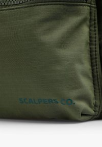 Primer plano de una tela color verde oliva con un patrón de cuadrícula y el logo azul "SCALPERS CO." impreso cerca del borde inferior.