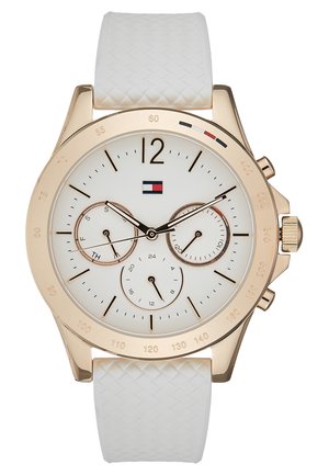 Gold-tone wristwatch with white textured strap, white dial, three subdials for day, 24-hour, and date, black and gold hands, and Tommy Hilfiger logo.