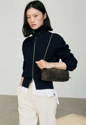 Femme portant un pull noir côtelé zippé, une blouse blanche et un pantalon crème, tenant un petit sac à bandoulière marron en daim.