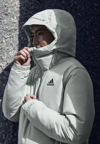 Lichtgrijze gewatteerde jas met een capuchon, voorzien van een verstelbare trekkoord, zwart logo en tekstdetail op de mouw, tegen een gestructureerde achtergrond.