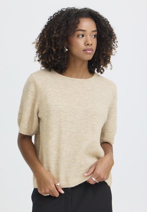 Fiatal nő göndör hajjal, bézs rövid ujjú pulóvert és fekete nadrágot visel, jobbra néz, kezei a dereka közelében.