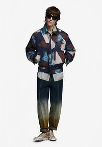 Veste en denim patchwork avec des motifs triangulaires en bleu, marron et crème. Jean dégradé foncé avec des ourlets effilochés et baskets blanches.