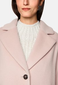 Helle rosa Wolljacke mit Reverskragen, einem einzelnen Knopfverschluss und einem strukturierten weißen Strickpullover darunter.