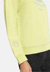 Limonkowa bluza z długimi rękawami, z niebieskimi kamieniami szlachetnymi w kształcie uśmiechniętej buźki na mankiecie. Tekst nadrukowany z przodu.