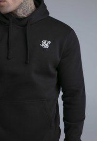 Uomo con felpa con cappuccio nera con logo bianco ricamato sul petto e tatuaggi visibili sul collo, su sfondo grigio uniforme.