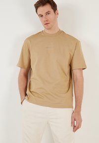 Tan kortærmet t-shirt lavet af blødt stof, med rund hals og en diskret broderet logo på brystet, iført med lyse bukser.