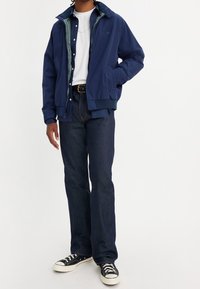 Veste bomber bleu marine avec une fermeture éclair, portée sur un t-shirt blanc, associée à un jean en denim foncé et à des baskets noires avec des semelles blanches.