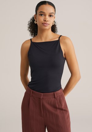 Fiatal nő göndör hajú, arany karika fülbevalóval, fekete ujjatlan felsőben és magas derekú vörösesbarna nadrágban, kezét hátul összekulcsolva áll.