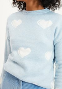 Jasnoniebieski sweter w białe wzory serc. Miękka faktura, okrągły dekolt, długie rękawy, ściągacze przy rękawach i na dole.
