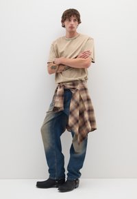 T-shirt beige a maniche corte, jeans azzurri chiari con zone scolorite, flanella a quadri marrone legata in vita, stivali neri alla caviglia. Look semplice e casual.