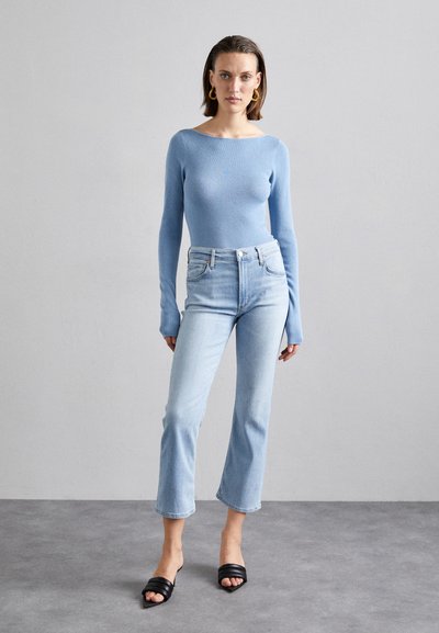 Vaaleansininen ribattu pitkähihainen paita yhdistettynä vaaleansinisiin leveälahkeisiin farkkuihin. Mallilla on jalassaan mustat kaksinauhalla varustetut liukuvaat. Yksinkertainen, moderni muotoilu.