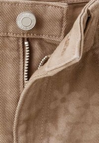 Pantaloni beige in cotone con chiusura a bottone metallico, cerniera e dettagli cuciti. Tessuto morbido, trama del tessuto visibile e variazioni di colore.