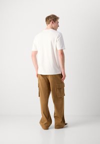 White short-sleeve t-shirt and brown cargo pants. Pants feature two side pockets and loose fit. Background is plain and light-colored.