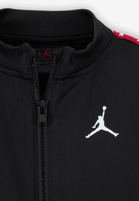 Schwarze Reißverschlussjacke aus glattem Stoff. Verfügt über einen hohen Kragen, ein weißes Jumpman-Logo auf der Brust und rote Akzente entlang der Schultern.
