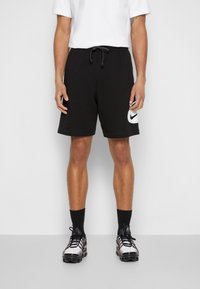 Shorts Nike noirs avec une taille élastique et un cordon de serrage, arborant un logo blanc proéminent sur le côté gauche. Portés avec des chaussettes et des chaussures noires.