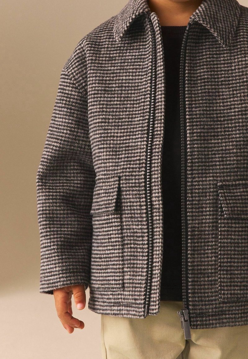Enfant portant une veste zippée à carreaux gris et noirs avec col, sur un t-shirt noir et un pantalon beige, sur un fond uni.
