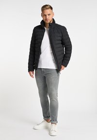 Schwarze Daunenjacke mit hohem Kragen, mit gesteppten Abschnitten. Getragen über einem weißen T-Shirt, kombiniert mit grauen Jeans und weißen Sneakers.
