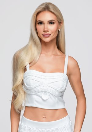 Blonde vrouw met lang haar die een witte cropped top met gevlochten details draagt en bijpassende hooggetailleerde broeken tegen een effen achtergrond.