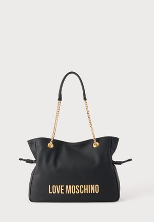 Must Moschino käekott kuldsete kettidega käepidemetega ja esiküljel logokirjaga, eksponeeritud lihtsal heledal taustal.