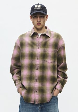 Jeune homme portant une chemise à carreaux rose et olive à boutons, jean bleu et casquette noire avec texte blanc, mains dans les poches, face à nous.