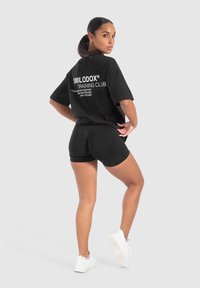 Sort oversized t-shirt med hvid tekst, kombineret med sorte shorts. Model vender væk, hvilket fremhæver et enkelt design og sporty stil. Hvide sneakers.