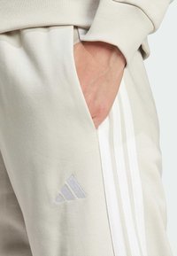 Beiga sweatpants med sidofickor, tre vita vertikala ränder och en ljusgrå broderad logotyp nära fickan. Mjuk textur.