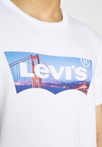 Valkoinen puuvillapaita, jossa on sininen ja oranssi graafinen kuva sillasta ja "Levi's" näkyvästi suurilla valkoisilla kirjaimilla.