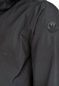 Svart vattentät jacka med en strukturerad textil, prydd med en rund svart logotyp på överarmen, som visar upp en stilren design och funktion.