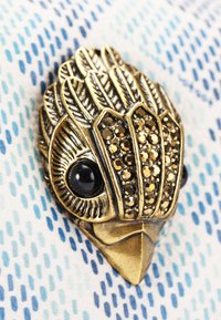 Bronzen broche in de vorm van een vogel, met zwarte stenen ogen en decoratieve details met glanzende gouden accenten, tegen een blauwe met patroon achtergrond.