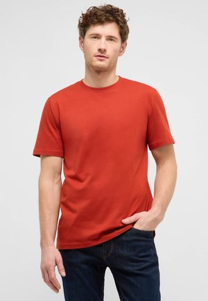 Man with curly hair wearing plain red short-sleeve t-shirt and dark blue jeans, standing with one hand in pocket against white background.